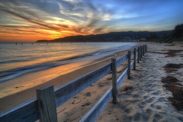 Sunrise over a beach with a wooden fence.  Sunrise hues paint the sky, gently lapping waves meet a sandy shore, and a simple wooden fence stretches into the distance