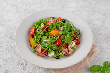 Salad with peaches, arugula, prosciutto, mozzarella cheese and cherry tomatoes with balsamic vinegar in a ceramic plate on a concrete background. Selective focus.
