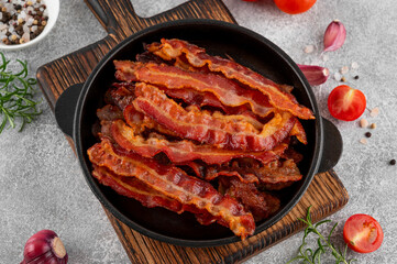 Fried slices of bacon for breakfast in a cast iron pan with tomatoes, garlic, rosemary and spices on a concrete background. Top view.
