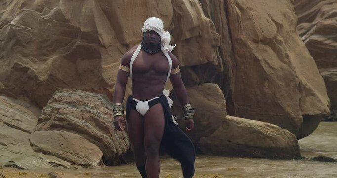 A dark-skinned, muscular male cosplayer poses on the beach.