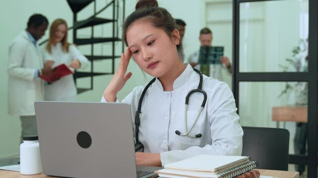 Exhausted healthcare professional experiencing burnout, rubbing temples while sitting at workstation with medical documents and computer in clinical setting. Asian chinese tired doctor or nurse 