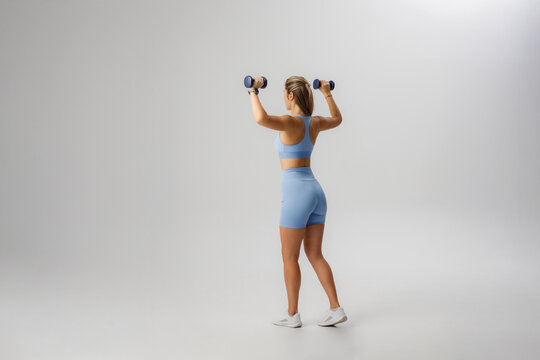 Fit woman exercises with dumbbells in stylish blue athletic wear indoors during bright lighting session