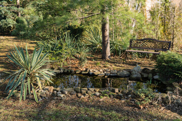 Serene garden scene with small stone-lined pond surrounded by lush greenery, including tall plants and trees. A wooden bench sits nearby, inviting relaxation amidst nature.