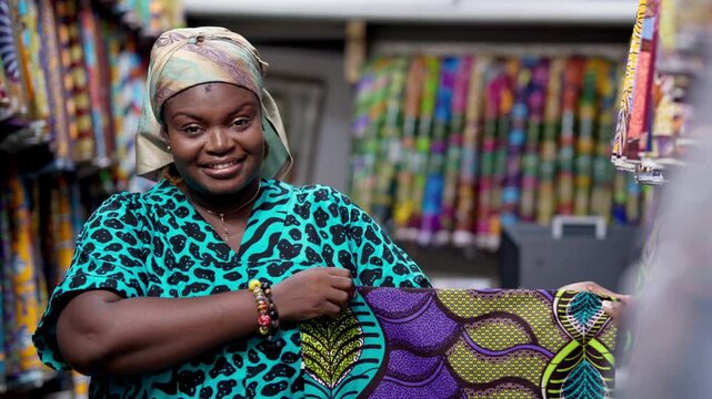 Smiling African woman, successful small business owner of an African Print, Ankara fabric shop proudly displays piece of textiles cloth.