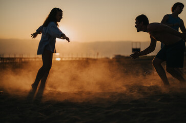 A lively scene of friends interacting playfully on a serene beach at sunset, with dust clouds...