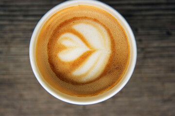 Top down view of a paper coffee  cup, placed on a rustic wooden table, filled with a creamy latte featuring heart and flower shaped latte art
