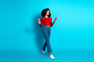 Cheerful young woman in red top holding coffee and phone against blue background embracing vibrant casual lifestyle