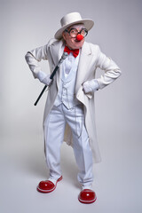 An elderly clown in a white coat and hat with walking stick with a cocky expression on his face and his hands on hips.