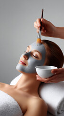 A young woman is lying down in a spa salon, getting a facial mask applied with a brush on her face
