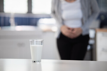 Allergy dairy food lactose intolerance allergen health care illness concept. Close up a glass of milk with blurred Asian woman holding stomach belly feel pain digestive unhappy after drink a cow milk.