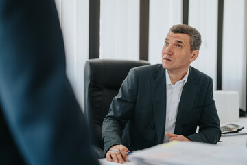 A businessman in a suit is sitting at his desk, attentively focusing on a conversation with a...