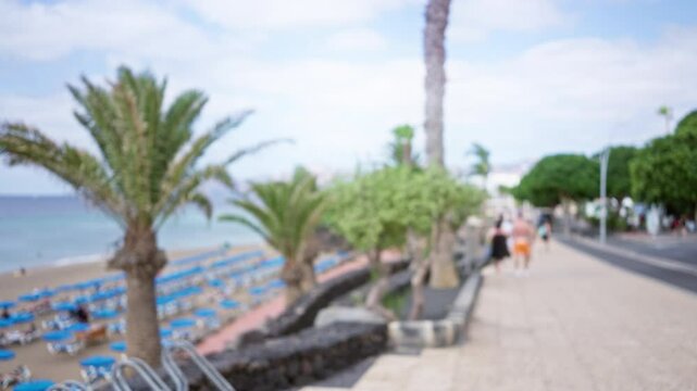 Defocused beach scene with blue loungers, palm trees, and blurred people walking on a beachside path on a sunny day