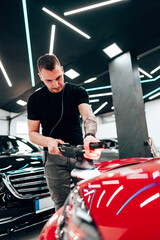 Professional car detailer using an orbital polisher to polish the hood of a red car, ensuring a...
