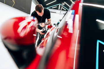 Professional car detailer using an orbital polisher to polish the hood of a red car, ensuring a...