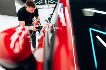 Professional car detailer using an orbital polisher to polish the hood of a red car, ensuring a...