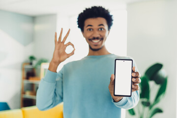 Young man in casual sweater indicating positive gesture while showcasing smartphone at home