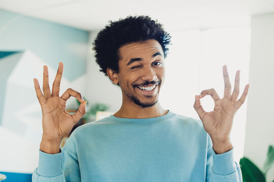 Happy young man indoors with a cheerful smile and relaxed posture showing an okay sign in a casual blue sweatshirt in natural daylight.