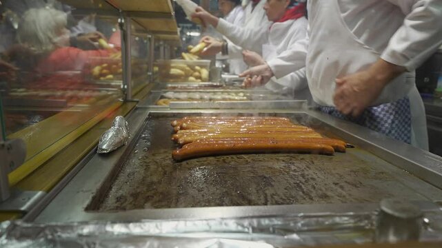 Close-up of a cooks hand frying Bavarian sausages in the evening at a fair in Germany, Munich. Bratwurst on barbecue grill. Street food. Outdoor food at the local market of the traditional festival. 
