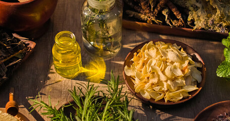 Top view of white dried flower petals placed on a wooden plate. On the table are many herbs such as...
