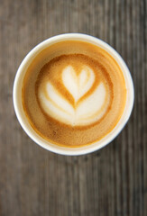 Top down view of a paper coffee  cup, placed on a rustic wooden table, filled with a creamy latte featuring heart and flower shaped latte art