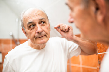 Portrait of senior man removing ear hair with tweezers while standing in front of bathroom mirror. 