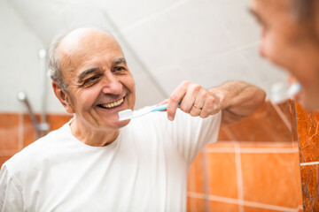 Portrait of senior man with bright smile looking at his reflection in mirror while brushing his teeth in bathroom. 