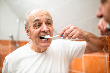 Portrait of senior man with gray hair brushing his teeth in front of the bathroom mirror.