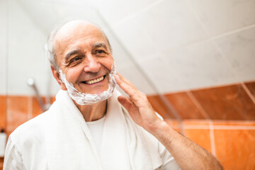 Happy senior man with wrinkled skin applying foam on his face and preparing for shaving while standing in front of bathroom mirror.