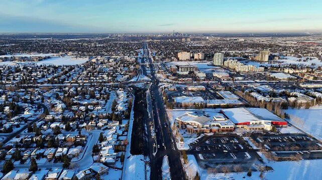 Aerial drone shot of a snowy suburban area in Edmonton, Alberta, Canada, with a wide highway leading toward the downtown skyline in the background on a clear winter day. Cars moving on the road, subur