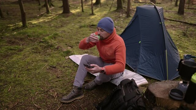 Male hiker drinking tea and surfing Internet on smartphone in tent camp during hiking in fall in woods. Backpacker makes route in map in phone and drinks hot drink from cup in background of tent. 