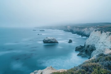 Misty coastal landscape, tranquil ocean meeting cliffs. Coastal rocks and fog create a serene scene. Ocean waves gently lap against the shore