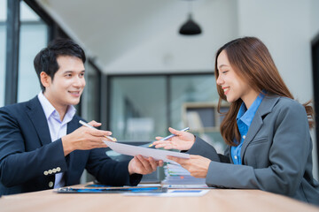 Two Asian business professionals exchanging documents and smiling during a meeting in a modern office, highlighting teamwork, collaboration, and positive communication