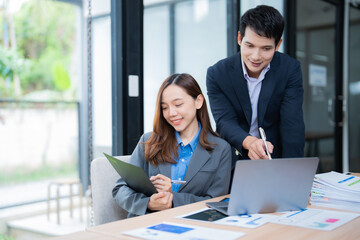 Two young professionals collaborating on a project, analyzing data on a laptop and reviewing documents in a modern office, exemplifying teamwork and focused effort