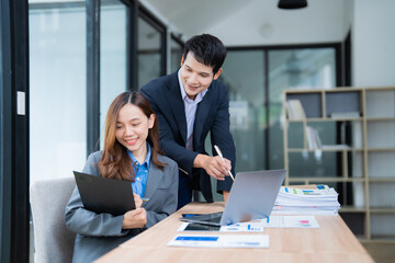 Two asian businesspeople are discussing and working together using laptop and documents, smiling and collaborating on a project in a modern office