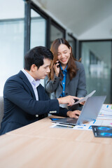 Two asian businesspeople are working together in the office, using a laptop and analyzing documents, while the woman is talking on the phone