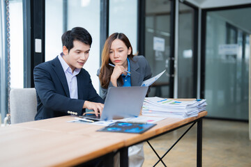Two asian businesspeople are working together in a modern office, using a laptop to analyze financial statistics displayed on the screen while examining reports and paperwork