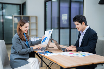 Two Asian business professionals engaged in a discussion about financial charts while seated at a modern office desk, utilizing laptops and reviewing paper documents for analysis