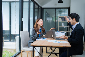 Two asian businesspeople are discussing and working together using laptop computer and analyzing financial chart documents while sitting at desk in modern office workplace