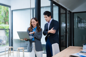 Two asian businesspeople discussing over laptop, smiling businesswoman holding laptop and sharing information with businessman in modern office
