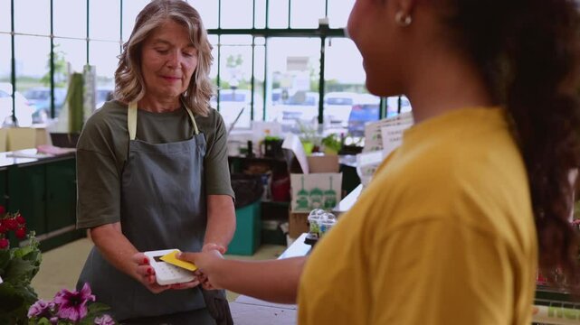 Friendly florist helping customer complete contactless payment using credit card at shop counter with flowers and natural light surrounding interaction
