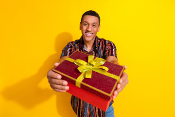 Smiling young man holding a gift box with a yellow ribbon on a vibrant yellow background,...