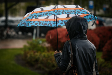 A woman dressed in a two-tone raincoat and jeans walks on a wet brick path holding a vibrant, patterned umbrella, with spring greenery and urban buildings in the background.