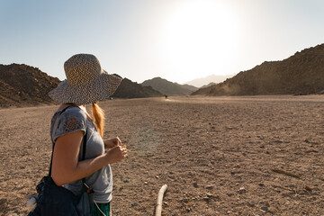 A woman in a wide-brimmed hat stands in a vast desert, gazing at the horizon.  The sun shines...
