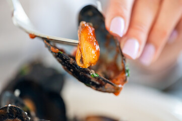 close-up of a hand holding a mussel shell with a fork, extracting cooked mussel meat covered in a...