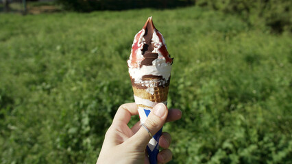 Hand holding chocolate and strawberry ice cream cone outdoors on a sunny day, with green grass in the background. Summer treat and refreshment.