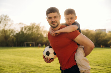 Boy is sitting, holding soccer ball. Father and son are having fun outdoors