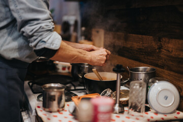 A person preparing food in a home kitchen, with pots, utensils, and tools organized on the counter, set against a wooden back splash, capturing the warmth of home cooking and culinary creativity.