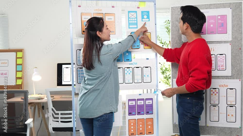 Poster two asian ux/ui designers arranging different versions of mobile app interfaces on a vertical whiteb