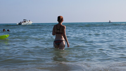 A young woman in a colorful bikini walks into the sea on a beautiful sunny day.  Boats are visible in the distance.