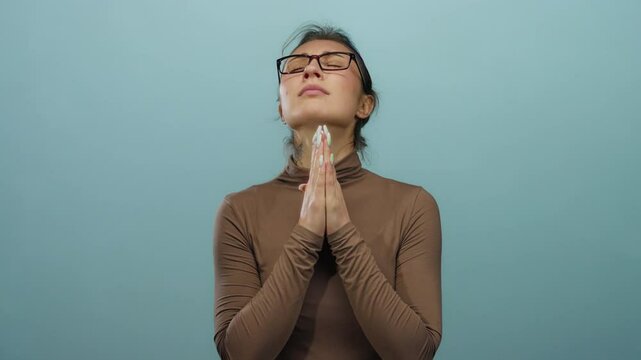 Young woman with glasses and a brown top praying with clasped hands against an isolated blue background wall